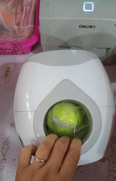 White electronic device with a green ball inside, on a pink surface with a Deli brand appliance in the background.