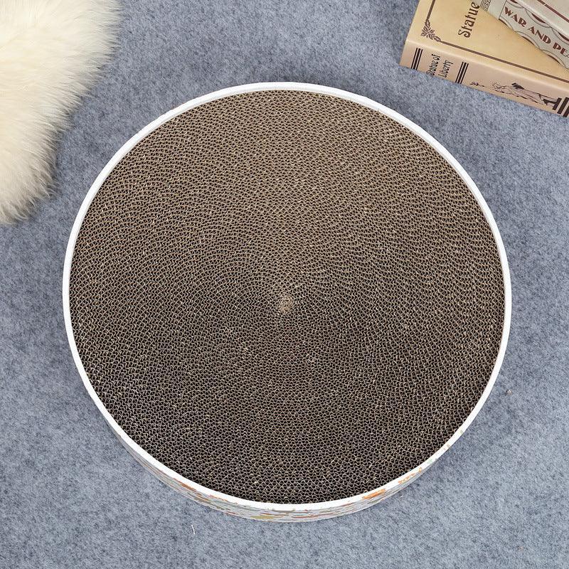Round cat scratcher on a gray surface with books and a fluffy object in the background